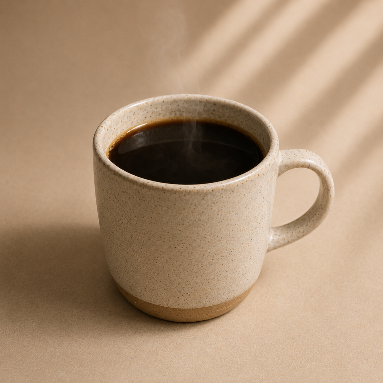 A speckled beige ceramic mug filled with black coffee, shown on a warm beige background with soft diagonal shadows.