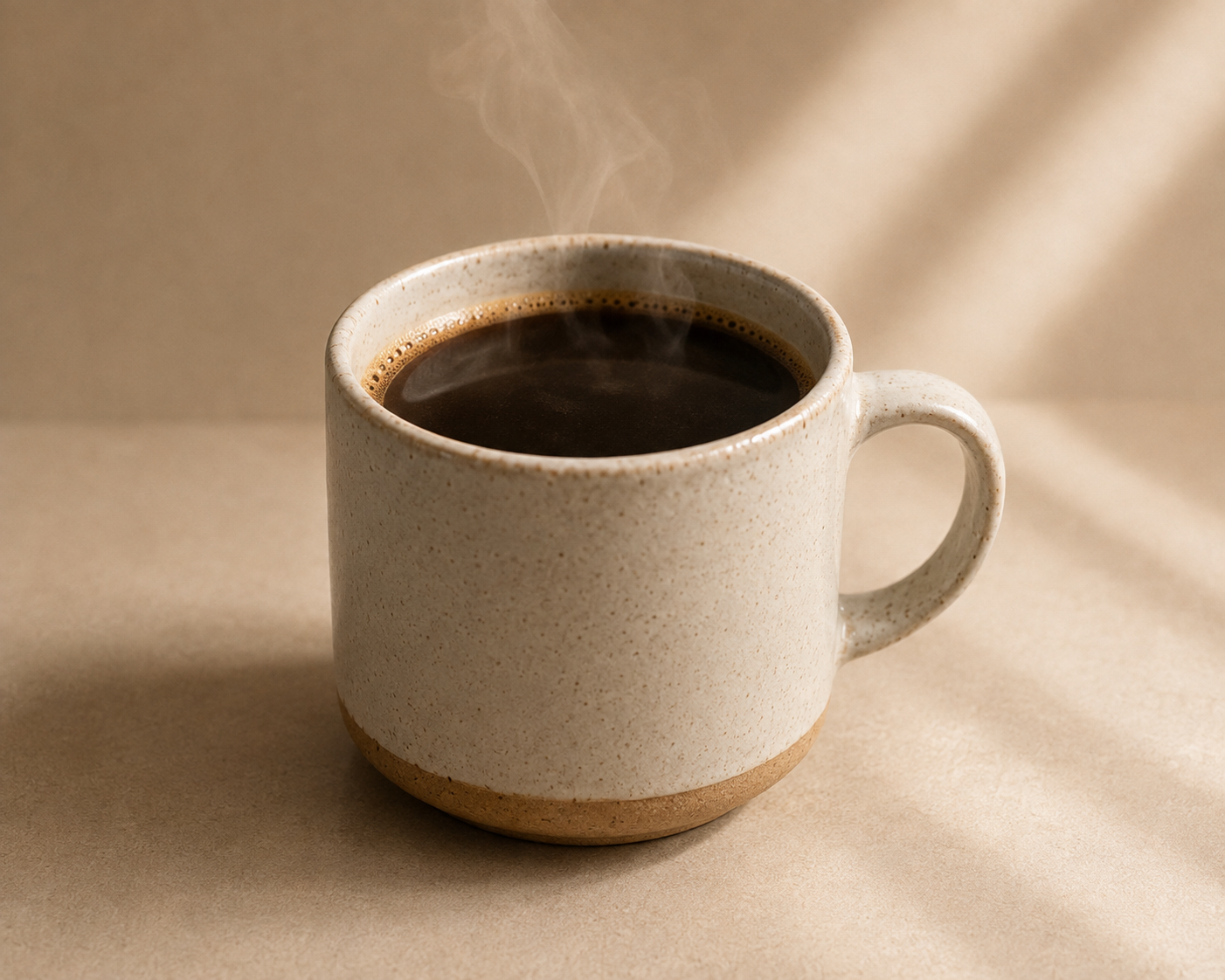 A speckled beige ceramic mug filled with black coffee, with steam rising, set against a warm beige background with soft diagonal shadows.