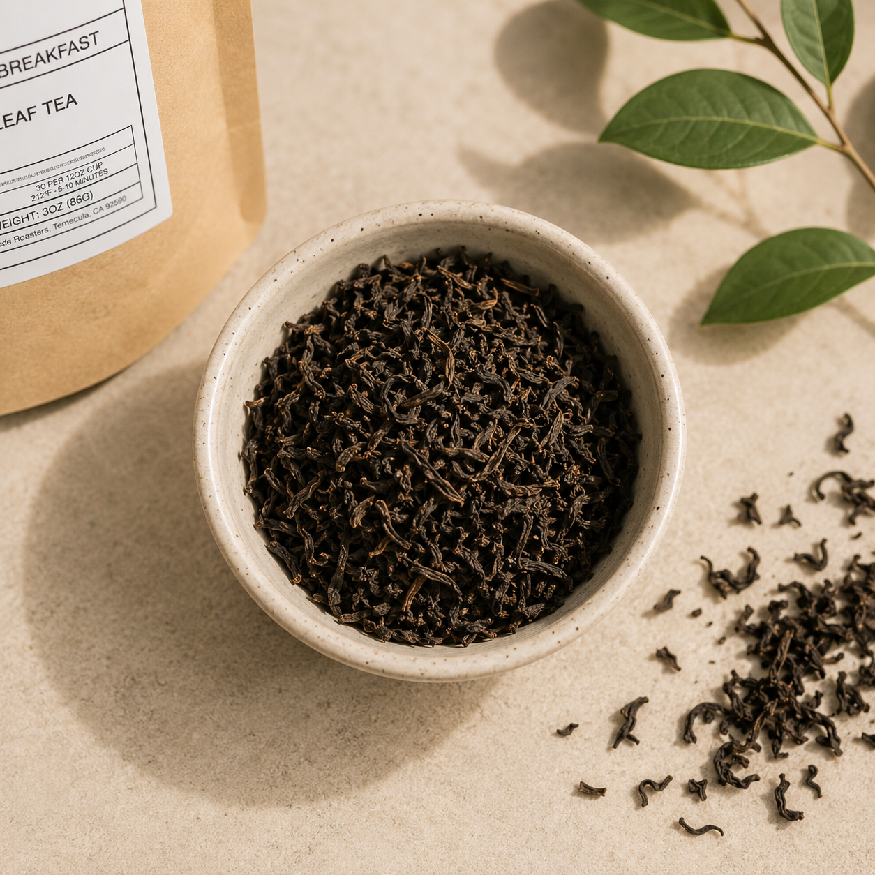 A ceramic bowl filled with dark loose-leaf tea sits on a beige surface, with scattered tea leaves nearby and part of a kraft paper tea bag package visible at the upper left. Green leaves appear in the top right corner.