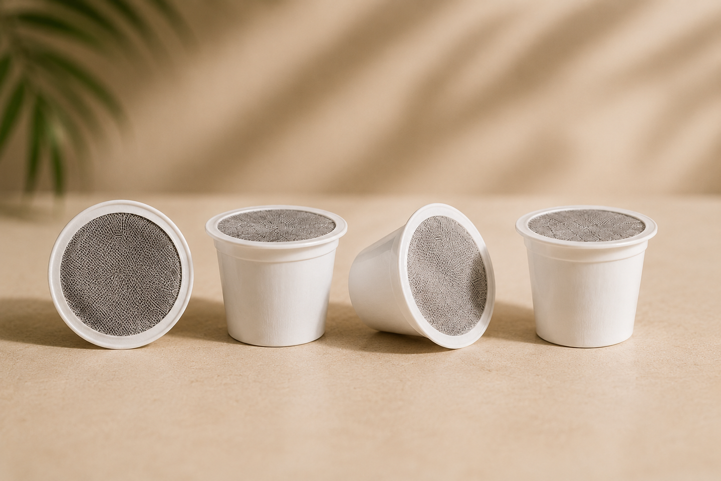 Four reusable white coffee pods or cup lids arranged on a beige surface, with a softly blurred warm background and a hint of greenery in the upper left.