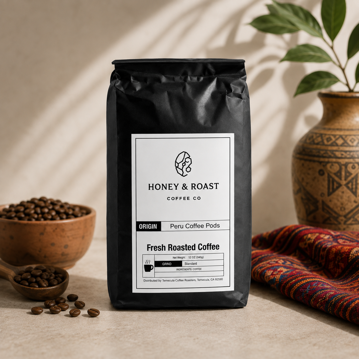 A black bag of Honey & Roast Coffee Co. Fresh Roasted Coffee stands on a beige surface with coffee beans, a wooden bowl, and a patterned textile nearby, under warm natural-looking shadows.
