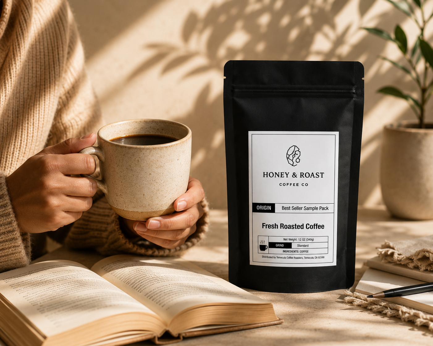 A person in a beige sweater holds a ceramic mug of coffee beside a black stand-up pouch labeled Honey & Roast Coffee Co. The scene is styled on a tabletop with an open book, sunlight shadows, and a small plant in the background.
