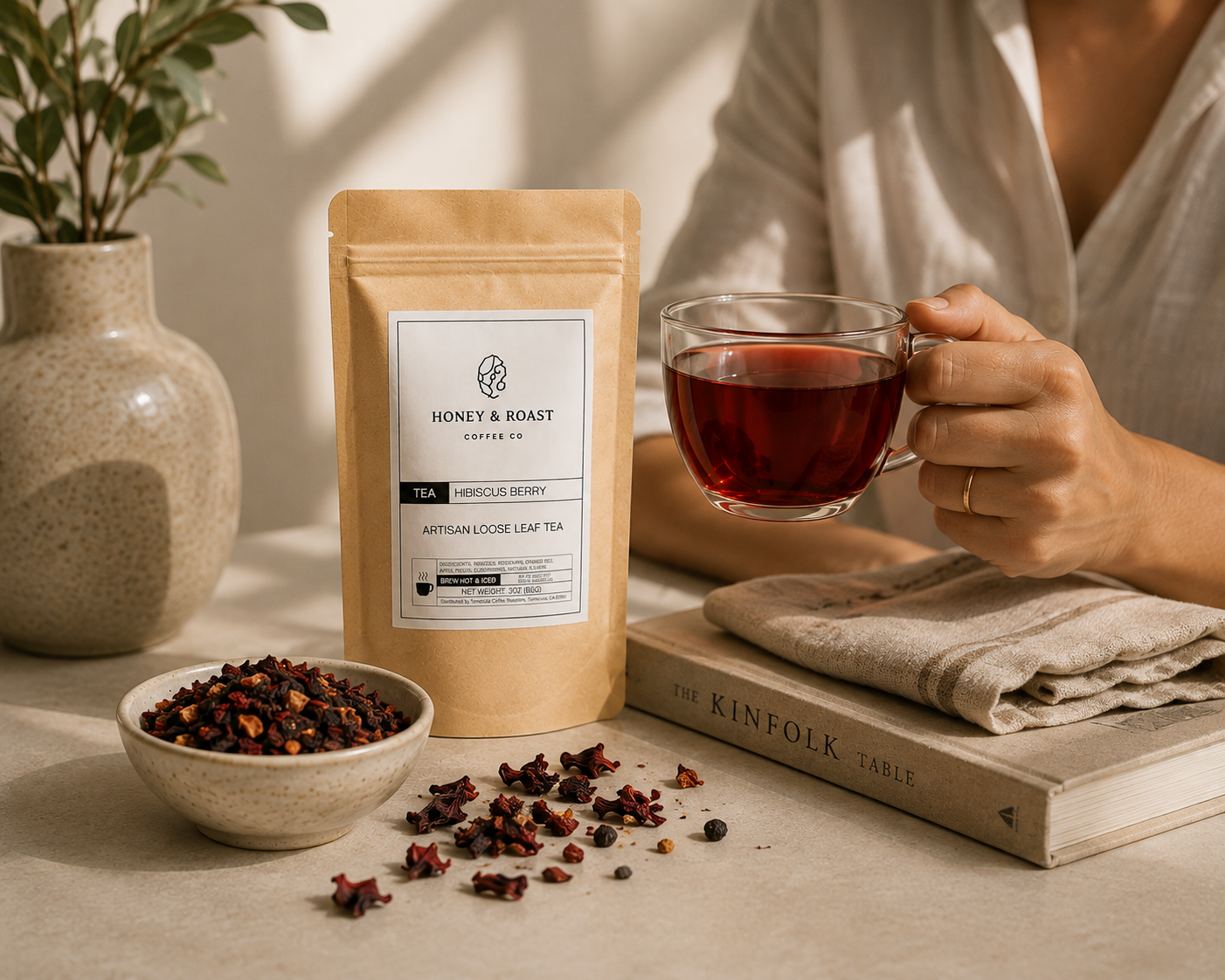 A stand-up pouch of Honey & Roast British Cosine Leaf Tea stands on a table beside a small bowl of loose tea, with a person holding a glass of the brewed tea in a warm, styled interior scene.