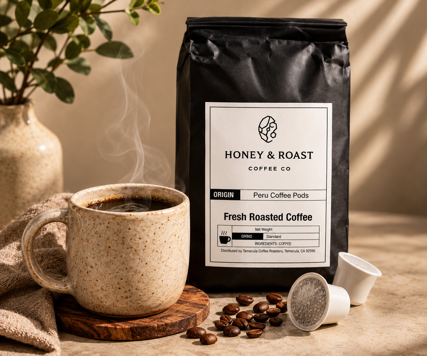 A black bag of Honey & Roast Coffee Co. Fresh Roasted Coffee stands on a tabletop beside a speckled ceramic mug filled with hot coffee, loose coffee beans, two coffee pods, and a plant in a vase.