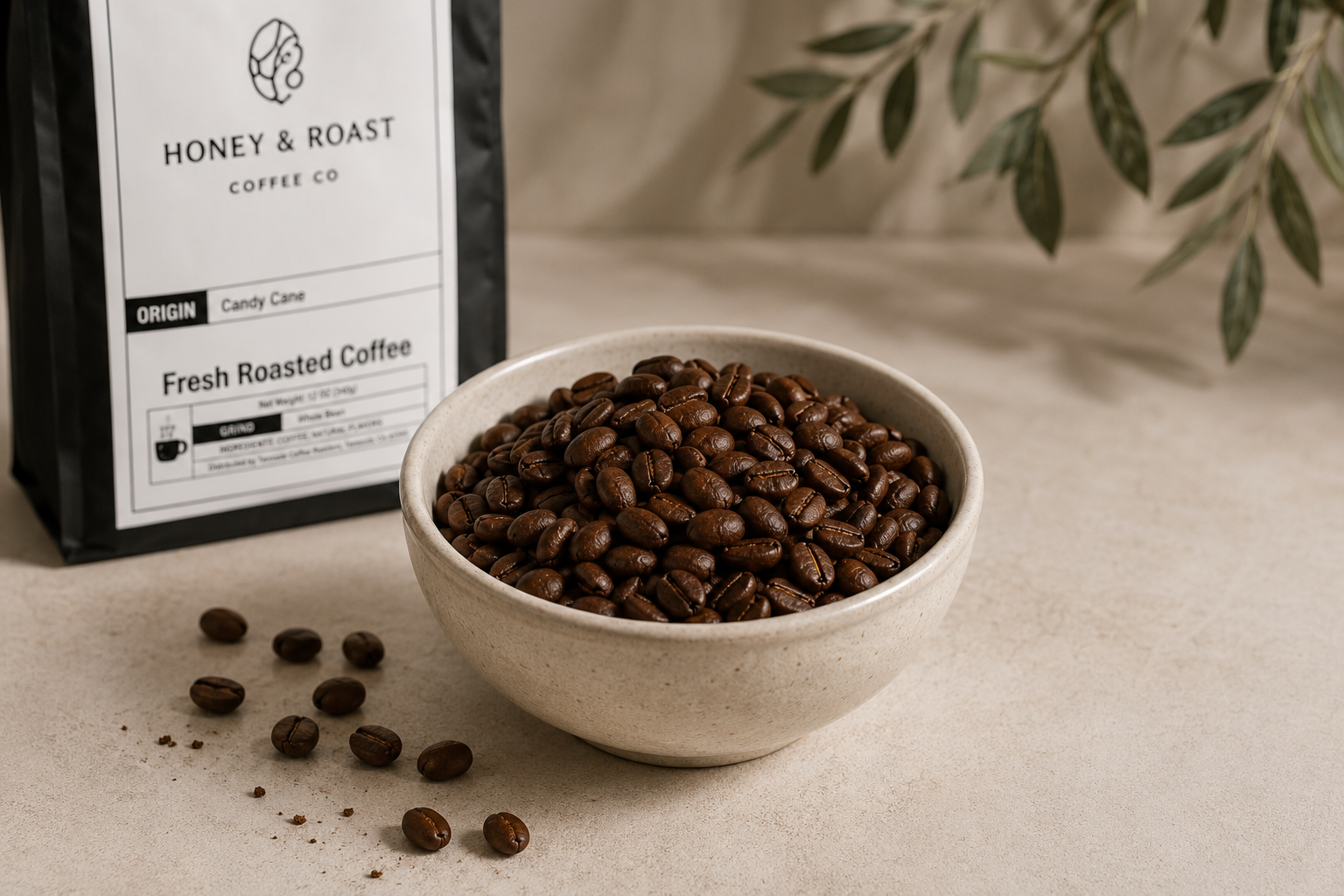 A bag of Honey & Roast Coffee Co. Fresh Roasted Coffee stands next to a ceramic bowl filled with dark roasted coffee beans, with a few beans scattered on a neutral beige surface and soft olive branches in the background.