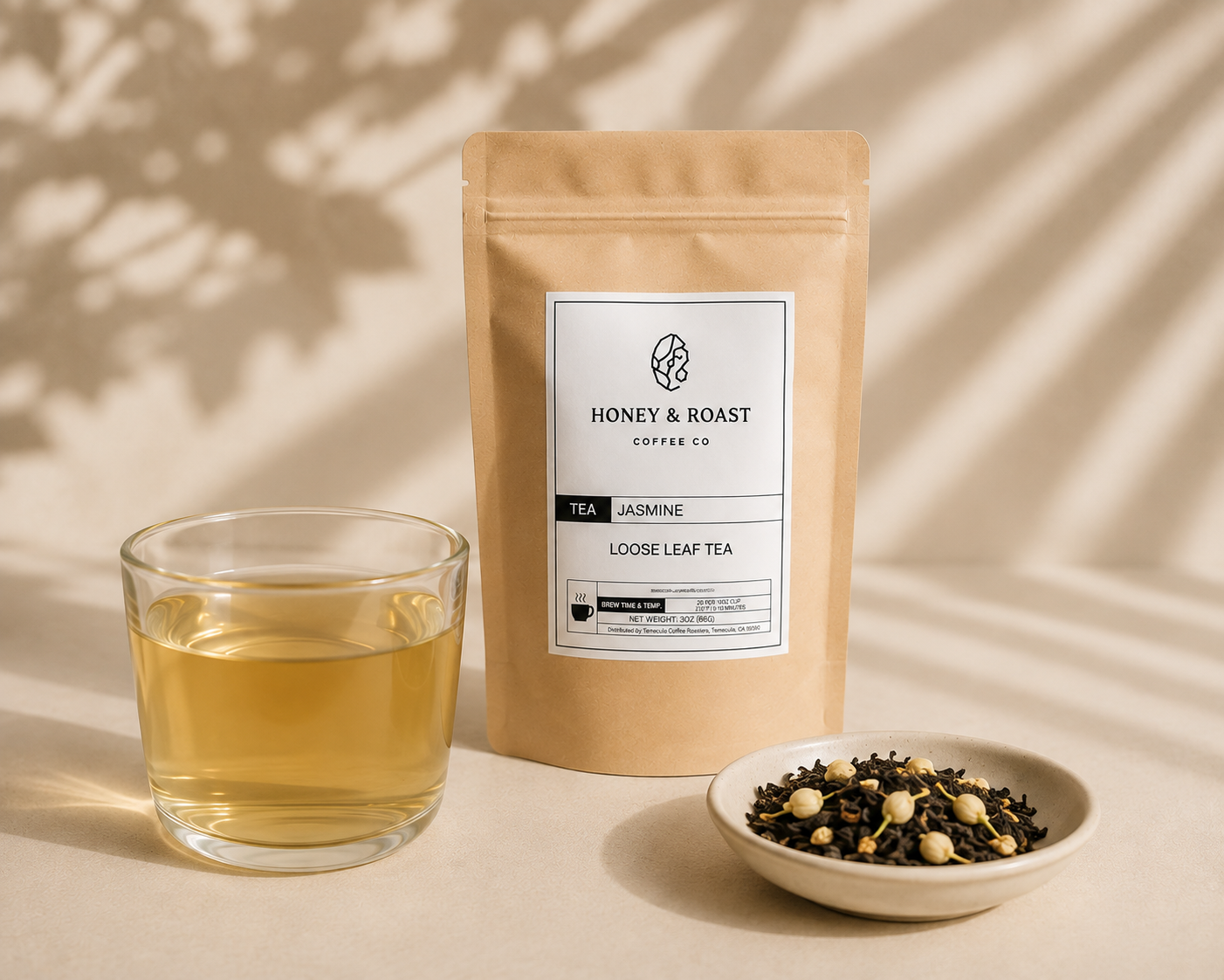 A kraft paper pouch of Honey & Roast Coffee Co. Jasmine Loose Leaf Tea stands next to a glass of brewed tea and a small dish of loose tea leaves on a warm beige surface with soft leaf shadows.