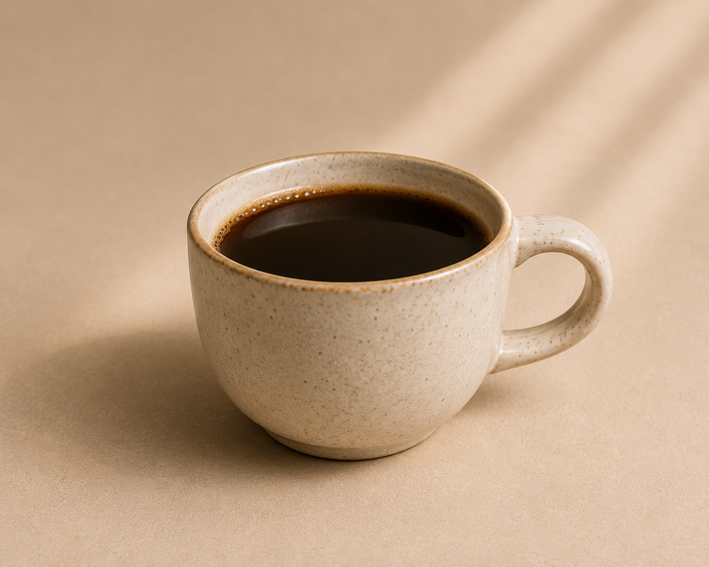 A speckled beige ceramic coffee cup filled with black coffee, set against a warm beige background with soft diagonal shadows.