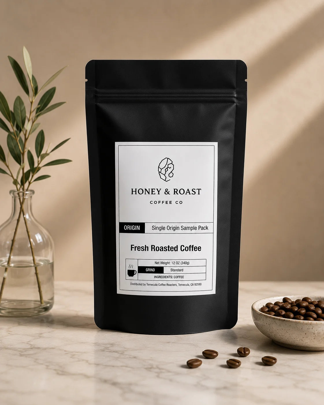 A black stand-up coffee bag labeled Honey & Roast Coffee Co. Fresh Roasted Coffee, placed on a light marble surface with a glass vase of greenery on the left and a small bowl of coffee beans on the right, against a warm beige wall with soft shadows.