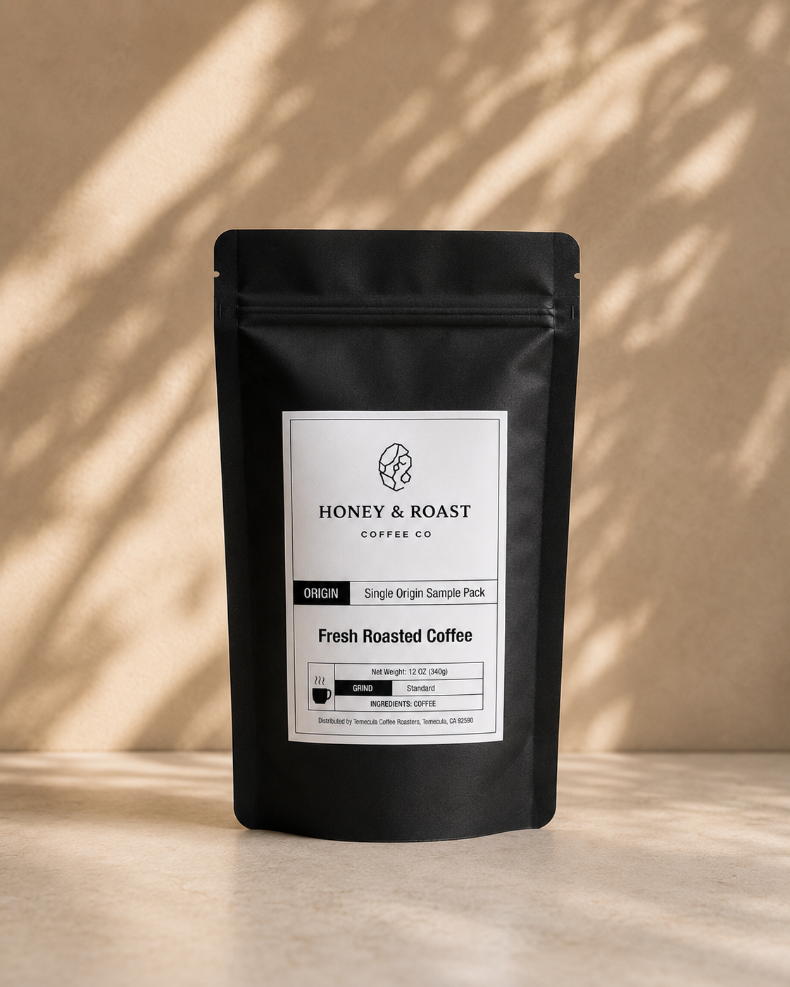 A black stand-up pouch of Honey & Roast Coffee Co. Fresh Roasted Coffee, featuring a white label, set against a warm beige background with soft leaf shadows.