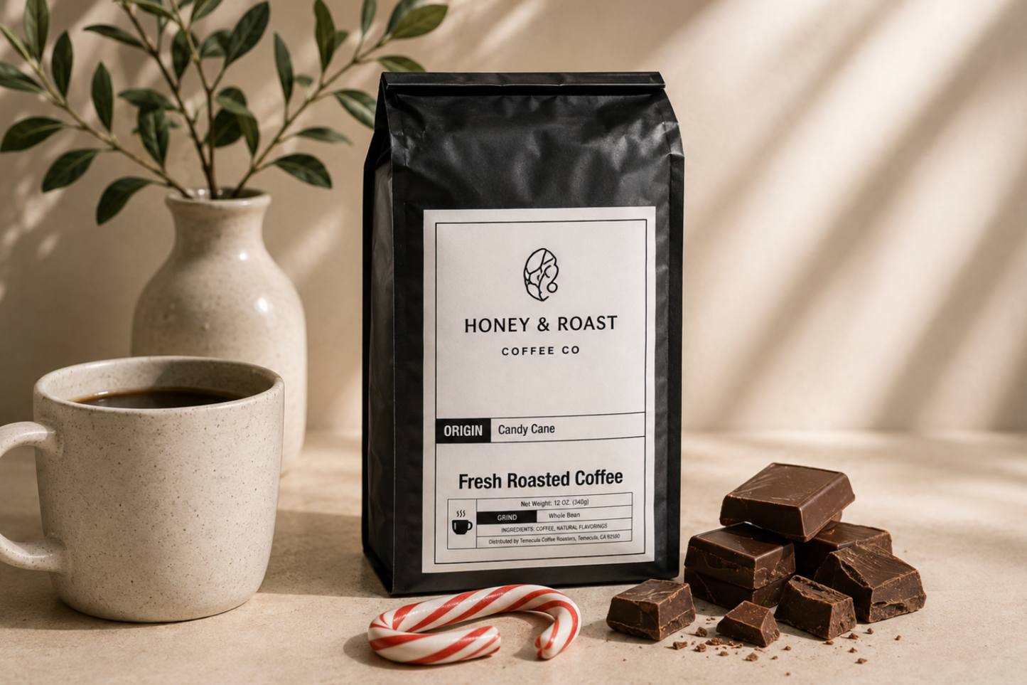 A black bag of Honey & Roast Coffee Co. sits on a tabletop beside a white mug, a small vase with leaves, candy canes, and chocolate pieces.