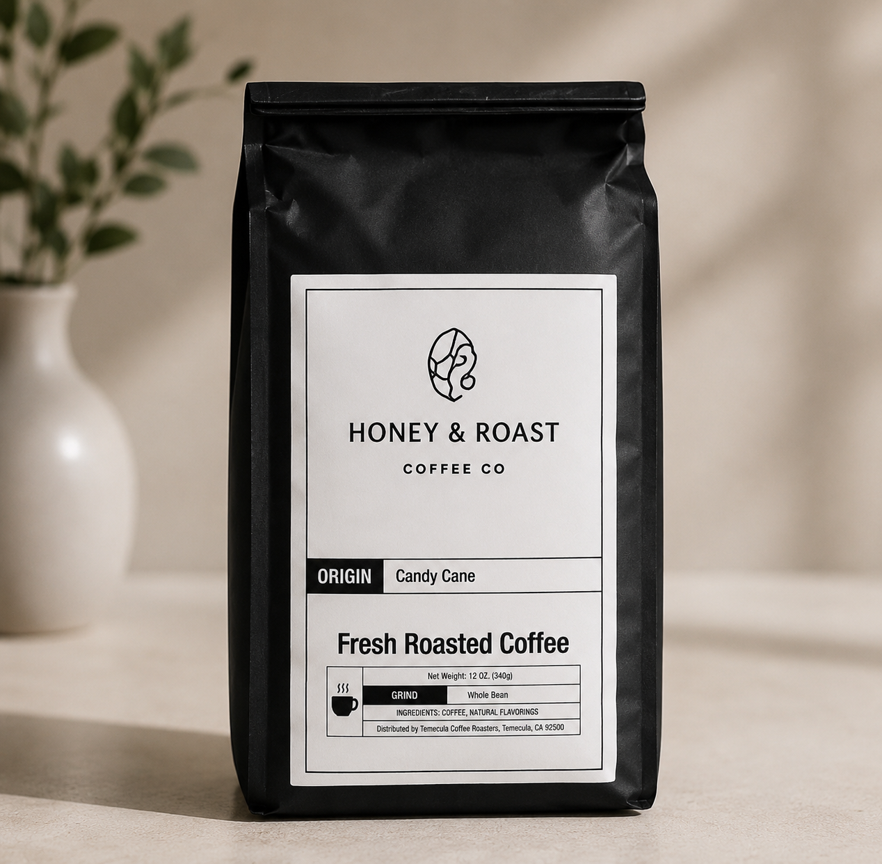 A black bag of Honey & Roast Coffee Co. Fresh Roasted Coffee, Candy Cane origin, standing on a light beige surface with a soft-focus background showing a white vase with green leaves.